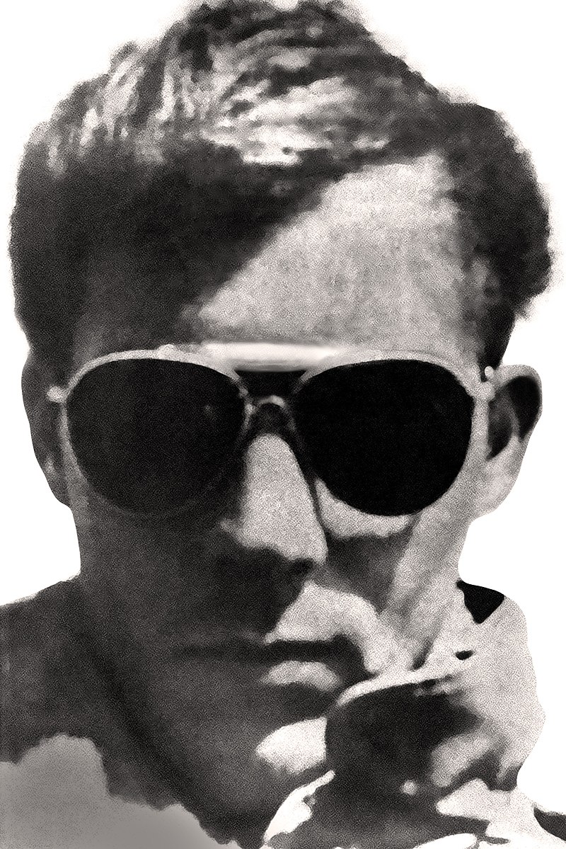 Photograph showing just the head of a man with a serious expression, aviator sunglasses, a full head of medium-short hair, and a visible collar of a leather jacket