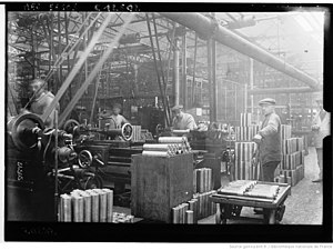 Photographie en noir et blanc d'une table couverte d'obus avec des ouvriers en arrière-plan.