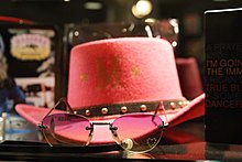 Image depicting a pink cowboy hat with a gold M amid two small gold stars. In front of the hat, pink sunglasses also appear.