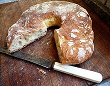 A fotografia representa uma coroa de pão.  O quarto retirado mostra um miolo com bolhas irregulares sob uma crosta dourada e enfarinhada.  O pão repousa sobre uma tábua de pão de madeira com uma faca dentada com cabo de plástico branco.