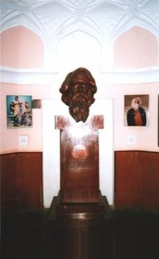 A bronze bust of a middle-aged and forward-gazing bearded man supported on a tall rectangular wooden pedestal above a larger plinth set amidst a small ornate octagonal museum room with pink walls and wooden panelling; flanking the bust on the wall behind are two paintings of Tagore: to the left, a costumed youth acting a drama scene; to the right, a portrait showing an aged man with a large white beard clad in black and red robes.