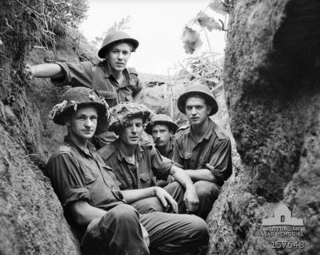 Five young Caucasian men in uniform wearing helmets sit below ground in a trench, facing the camera. The trench is central to the photograph and runs away from it, with the edges of the earthworks on each side. In the background, the skyline can be seen to the rear of the men, framed by the edges of the trench, which are covered in roots, grass, and other vegetation.