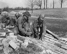 Five workmen. One is holding a shovel, while the other four are laying bricks to form a drainage ditch along the side of a road.