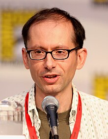 A closeup of a man in front of a microphone. He has a receding hairline and wears dark-framed glasses.