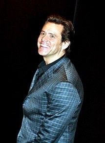 A Caucasian with dark brown hair and brown eyes, in front of a black background. He is wearing a chequered blue blazer and black shirt