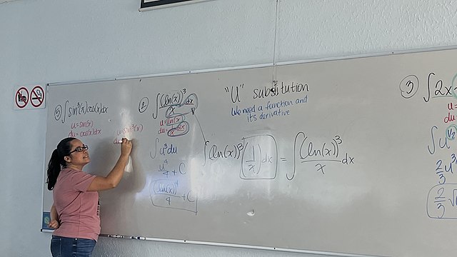 Maestra de cálculo escribiendo en una pizarra blanca sobre la cual desarrolla una explicación en clase.