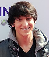 A head shot of a teenage boy with brown, straight hair, wearing a grey, hooded jacket. He is posing at a press event and smiling, looking directly towards the camera. Behind him are hoarding signs with names of commercial sponsors.