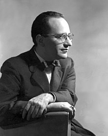 Murray Rothbard wearing glasses, a suit and a bow-tie and sat on an armchair, looking rightwards