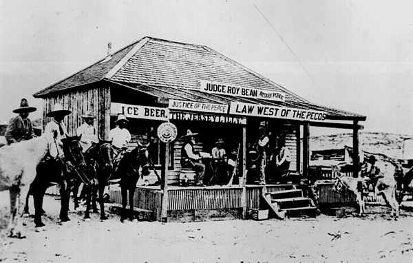 Roy Bean holding court while sitting on a barrel and holding his law book at his Jersey Lilly Saloon, 1900s