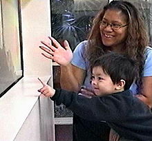 A 3-year-old child is points to something offscreen in an aquarium while a therapist smiles and looks where the child is pointing.