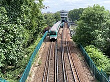 Foto af et metrotog set forfra på en overliggende jernbanelinje foret med buskede træer.