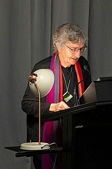A smiling elderly woman with short grey hair and glasses, her face lit by a lamp, reads from a podium