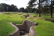 Curved trench lines, preserved in concrete, are surrounded by shell craters that are now covered in grass. In the immediate foreground, a small half-destroyer piece of artillery sits in a three-walled position that is off of the main trench line.