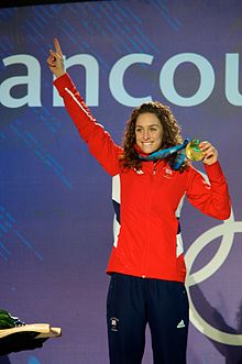 Eine Brünette mit langen, welligen Haaren lächelt oben auf einem Podium, als sie ihren rechten Arm hebt und mit der linken Hand eine Goldmedaille um den Hals hält. Sie trägt eine leuchtend rote Sportjacke und eine dunkelblaue Sporthose.