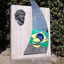 Foto de um monumento em memória de Senna no circuito da Catalunha em Montmeló formado por um retângulo de pedra com o vosage de Senna, seu capacete e a bandeira do Brasil.