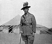 a man in uniform with shift and tie and Sam Browne belt, holding a walking stick and wearing a slouch hat with emu feathers. He is standing in front of a large tent.