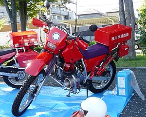 A pair of bright red off-road motorcycles, one partially obscured, both fitted with top boxes covered in Japanese writing. The motorcycles are parked on a blue plastic sheet laid on the ground