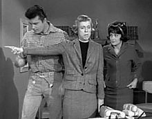 A black and white screenshot from the television series, The Beverly Hillbillies shows Max Baer, Jr. as Jethro, Nancy Kulp as Jane Hathaway, and Sharon Tate as Janet Trego, a secretary. Tate is wearing a business suit and a dark wig, and is watching Miss Hathaway