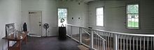 White pained interior of a room with wooden walls, three windows, and door. There is a wooden table at left and a white railing at left.