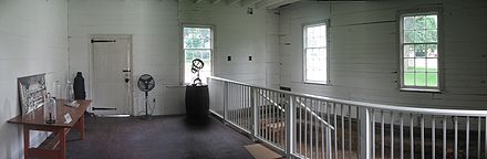 Intérieur peint en blanc d'une pièce avec des murs en bois, trois fenêtres et une porte.  Il y a une table en bois à gauche et une balustrade blanche à gauche.