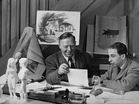 Photo de deux hommes attablés dans un studio, entourés de planches à dessin et de figurines, concentrés sur une feuille qu'ils examinent tous deux.
