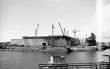 Campo lungo che mostra un cantiere ai margini del bacino del porto di Lorient.  Molte gru sono visibili sopra una massiccia struttura in cemento armato.