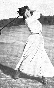 Wanita kulit putih di luar ruangan pada tahun 1901, yang mengenakan gaun putih panjang, mengayunkan klub golf.