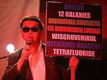 Chu holds a microphone and a sign. The sign reads "Bolton / 12 Galaxies / Duxbrowrenikal iconoclastics / Fox: Raxdrobrenicol coverage / Wisgrovenikul / Reticence nascent / Tetrafluoride