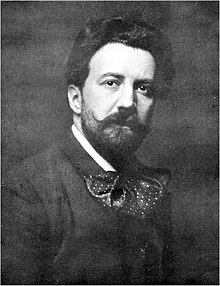 Head and shoulders picture of a young man with flowing medium length dark hair, a beard and moustache, and a huge floppy bow tie