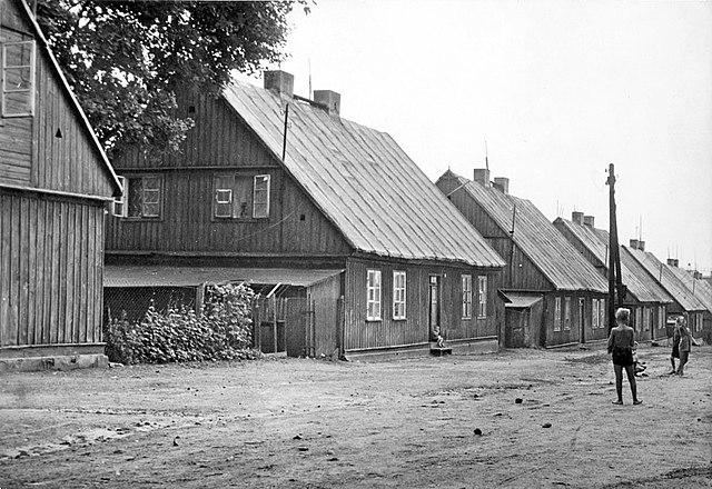 Fotografia czarno-biała. Gruntowa droga biegnąca w głąb zdjęcia, wzdłuż ulicy znajdują się identyczne, drewniane, dwukondygnacyjne, zwieńczone dwuspadowym dachem z dwoma kominami, budynki. Na środku dłuższej, skierowanej do ulicy, ścianie budynków znajdują się drzwi, a po obu ich stronach para okien, w ścianach prostopadłych do drogi para okien znajduje się na poddaszu. Na ulicy bawią się lekko ubrane dzieci.