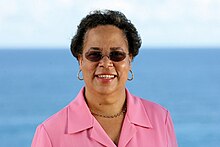 Joevana Charles. Porträt einer Frau mit hoher Stirn, großen Creolen und Sonnenbrille im ros hemd vor Meeresblauem Hintergrund.