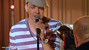 A white man in front of a camera holding a microphone with stand, wearing a purple and white striped shirt with beads at the neck, a beret on his head and a watch on his left hand.