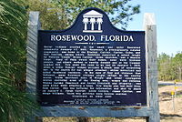 Una fotografía en color del frente de la placa de bronce en Rosewood junto a la carretera.