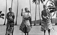 Three matai, the two older men bearing the symbols of orator chief status - the fue (flywhisk made of organic sennit rope with a wooden handle) over their left shoulder. The central elder holds the orator's wooden staff (to'oto'o) of office and wears an 'ie toga, fine matting. The other two men wear tapa cloth with patterned design Three Samoan chiefs including two orators - unknown photographer and date.jpg