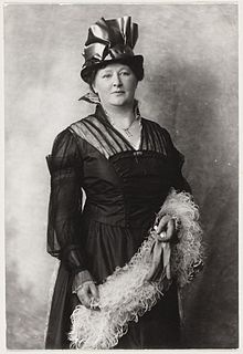 A white woman, standing, wearing a hat and a dark dress, and holding a fluffy scarf or stole