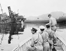Vista em preto e branco da frente de um barco a remo com 4 soldados.  Ao fundo, um navio naufragado.