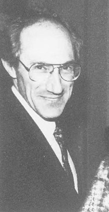 Photograph of a smiling, suit-wearing, middle-aged man with a receding hairline and wire-rimmed glasses