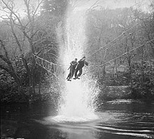 Two men crossing a rope bridge over water