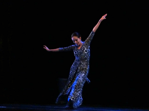 Na foto vemos Eliete Santos durante uma performance de dança. Fundo preto, a bailarina aparece de corpo inteiro congelando um movimento, braços acima da linha do ombro, pernas para o lado direito. Cabelo escuro preso, pele de tom médio, vestido longo em tons verde escuro.