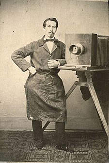 Jacob Hilsdorf, Fotograf mit Schürze an Kamera lehnend im Atelier, Sammlung Landesmuseum Koblenz.jpg