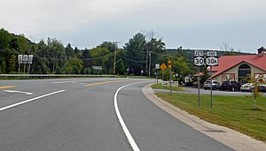 A broad stretch of curved paved road, with turn lanes and a double yellow line down the center, photographed from its side. At the right is a small building with a reddish cross-gabled roof; a sign on the road says it is the "Apple Barrel Café". In the front right are two black on white signs with the numbers "30" and "30A" on them