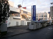 white 3-storey building; in the foreground is: a vertical blue banner with the Olympus logo