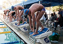 Olympian Ryan Lochte (near) standing on top of the wedged starting blocks. Each swimmer performs a preparatory isometric press by applying downward pressure onto their bent legs. This serves to preload the muscles and helps to make the subsequent dive more powerful. Ryan Lochte (4800088801).jpg