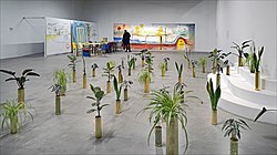 Depicts an installation by Sammy Baloji containing multiple plants in shiny containers spread out across the floor. The background piece an installation by Fabrice Hyber.