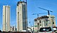 Ground level view of three towers; the one farthest to the left is complete, and has an all-white facade with balconies and light windows. The two buildings on the right are both under construction; the middle tower is nearly complete, with some concrete still exposed and a crane on the roof. The tower farthest to the right is also incomplete and has about 10 stories of framework constructed.