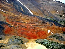 Orange, red and dark-colored rocks exposed on a rocky, lightly snow-covered mountain slope.