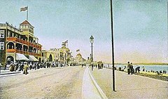 An early 20th century postcard view of a beach with a road running alongside. Across the road from the beach are some buildings and a wide sidewalk full of people. There are also people standing and walking on the beach.