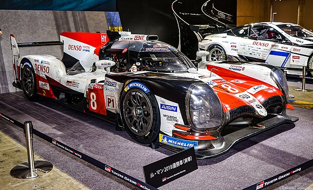 Side view of a red, white and black Toyota TS050 Hybrid enclosed in a space at an automobile show