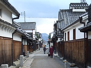 A narrow street lined by houses with a wooden lower part, a white upper storey and tile roofs.