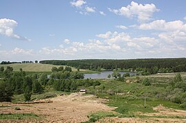 Paysage vallonné avec des forêts, champs et un petit lac artificiel au centre.